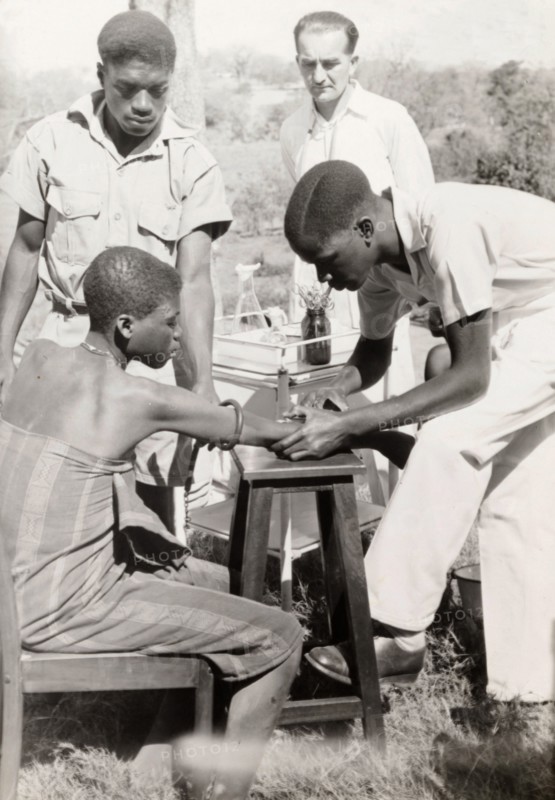 An African medic injects a patient. An African medic gives an injection to a female patient, while a European doctor stands by a medical trolley and observes. Probably Northern Rhodesia (Zambia), circa 1950. Zambia, Southern Africa, Africa.