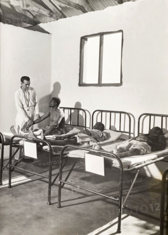 A European doctor examines African patients. A European doctor examines a patient as he sits on a hospital bed in a ward containing two other male patients. Probably Northern Rhodesia (Zambia), circa 1950. Zambia, Southern Africa, Africa.