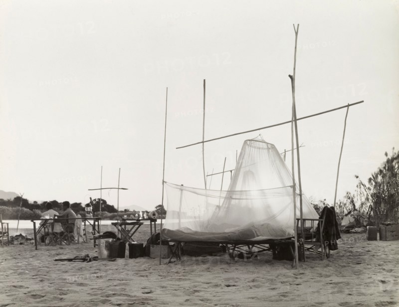 Camping on the banks of the Zambezi River. The camp site of a District Commissioner on the banks of the Zambezi River, where tents consisting of mosquito nets are strung up on a framework of wooden poles. Two men sit by a camp fire, watching dawn break over the river. Northern Rhodesia (Zambia), circa 1950. Zambia, Southern Africa, Africa.