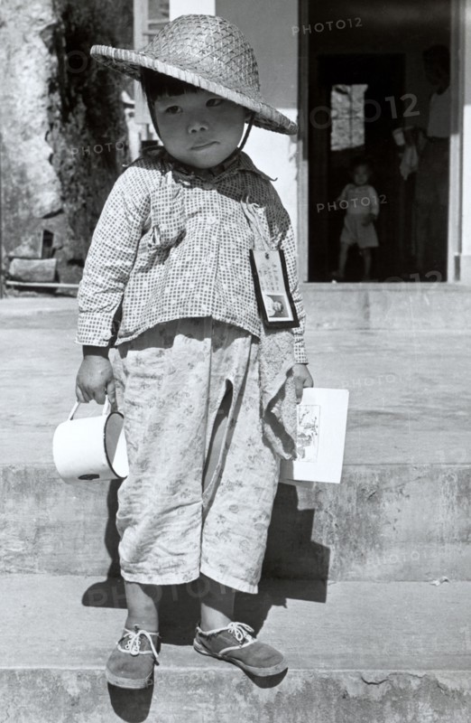 Refugee from China. A refugee boy from China stands on steps infront of an open doorway with an identity card and towel pinned to his shirt. He wears a straw hat and holds a tin cup in one hand. Hong Kong, People's Republic of China, 1963. Hong Kong, Hong Kong, China, People's Republic of, Eastern Asia, Asia.