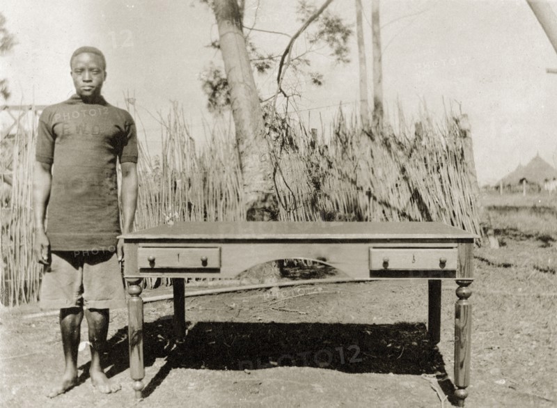Cabinetmaker with desk. A Public Works Department employee, identified in the the caption as 'Witaba Wa Sitamani', stands beside a desk with side drawers and turned legs that he has made. He wears a government uniform that displays the initials 'PWD' on the chest. British East Africa (Kenya), March 1916. Kenya, Eastern Africa, Africa.