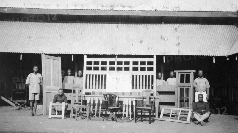 Cabinetmakers from the Public Works Department. Local employees of the Kenyan Public Works Department pose beside an array of doors, window frames, chairs and other wooden furniture constructed by them under the supervision of Charles Bungey, a training officer with the PWD. Nakuru, Kenya, 1924. Nakuru, Rift Valley, Kenya, Eastern Africa, Africa.