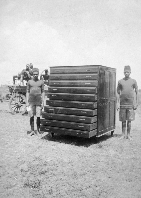 African cabinetmakers. Two young African men stand either side of a large map cabinet they have made as employees of the Public Works Department. Both men wear government uniforms and are identified in the caption as 'Makongoro wa Chancoro (sic) na Juma Wasila'. British East Africa (Kenya), 1914. Kenya, Eastern Africa, Africa.
