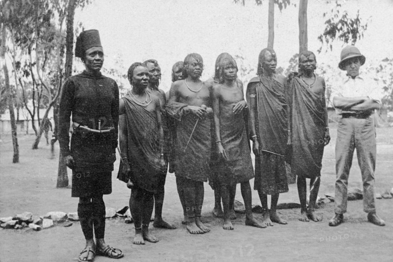 Raw recruits. A European officer and an African soldier stand either side of a small group of traditionally dressed African men, all new recruits for the King's African Rifles. British East Africa (Kenya), circa 1916. Kenya, Eastern Africa, Africa.
