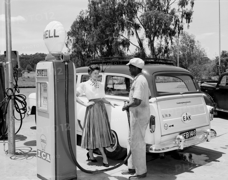 Daphne Dale fills up with Shell. Promotional shot of Daphne Dale, posing for the camera beside a car at a roadside petrol station. An African workman faces her, holding the hose of a Shell petrol pump. Kenya, 11 November 1955. Kenya, Eastern Africa, Africa.