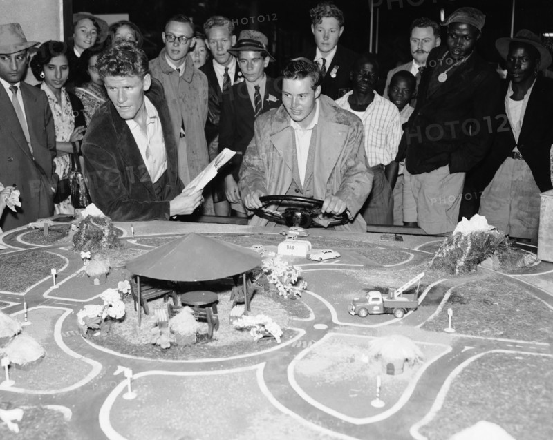 Driving game. A mixed race crowd gathers around a detailed miniature road model at the Motor Sports Club stand at the Royal Show. An European man holds a steering wheel and appears to be controlling the movement of the model cars. Nairobi, Kenya, 28 September-1 October 1955. Nairobi, Nairobi Area, Kenya, Eastern Africa, Africa.