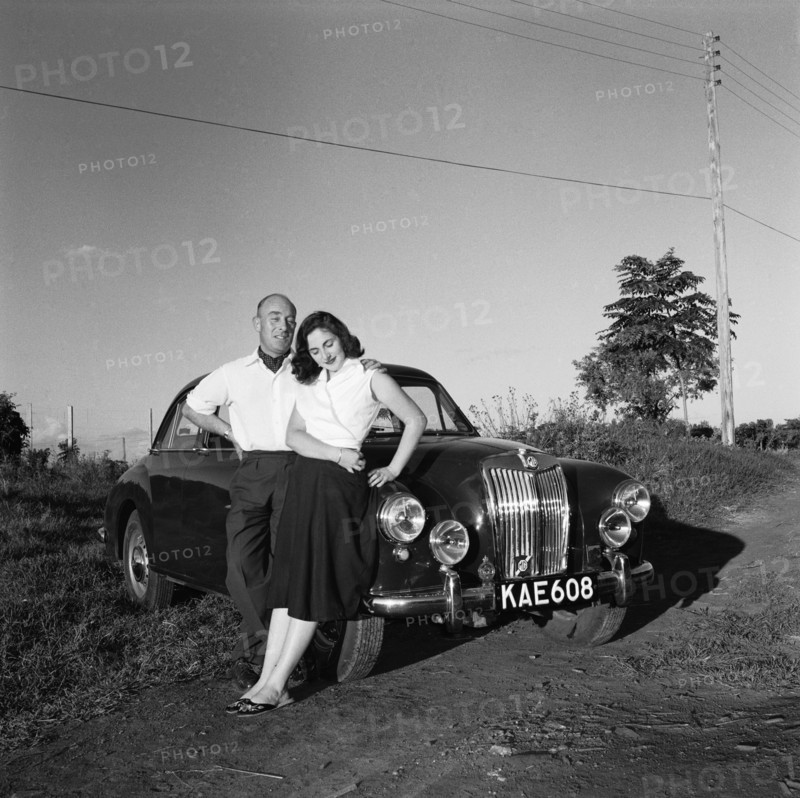 Sue and John Paton. Sue and John Paton relax, leaning back against their MG car in the evening sunlight. She wears a wrapover top and embroidered Chinese-style slippers: he wears a cravat. Kenya, 24 October 1955. Kenya, Eastern Africa, Africa.