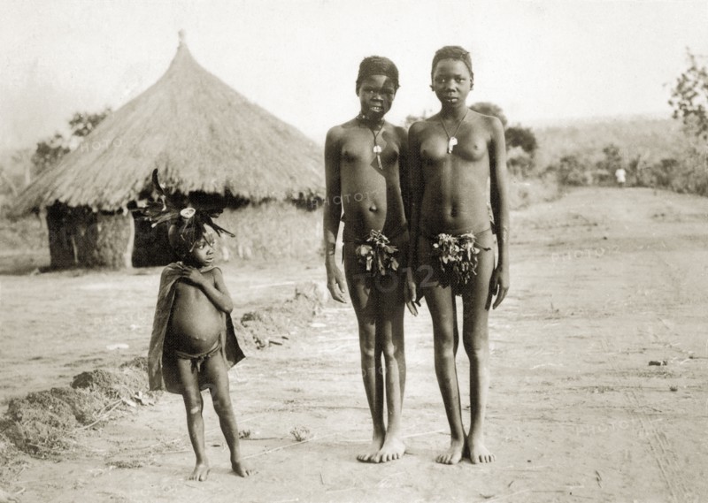 Balanda children on the Bo road. Three semi-naked Balanda children on the road to Bo town. The older girls wear necklaces and bunches of leaves that cover their genitalia: the young boy wears a cape and an elaborate headdress made from feathers. Sudan, circa 1930., West Bahr el Ghazal, Sudan, Eastern Africa, Africa.