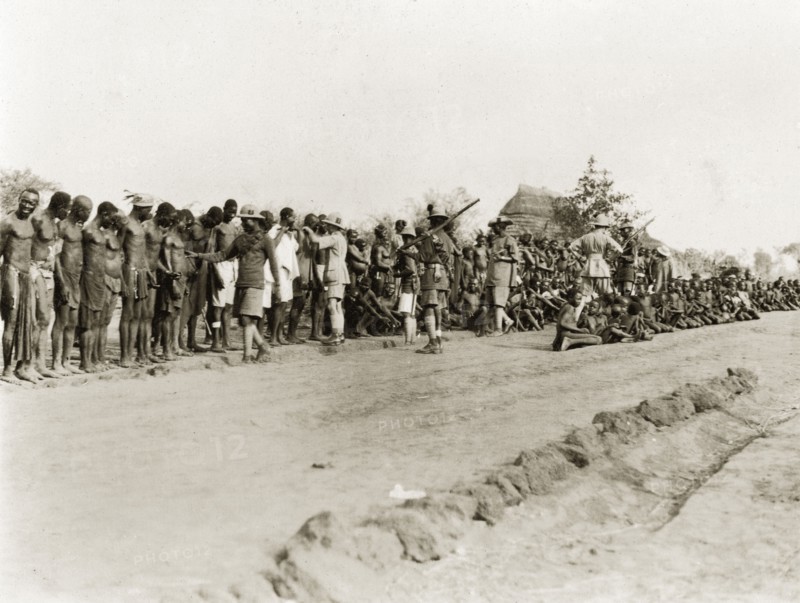 Sleeping sickness inspection. Sudanese men and boys stand and sit in a line on a roadside awaiting inspection for sleeping sickness by a senior medical officer. Armed African and European colonial officials in uniforms oversee the crowd. Sudan, February 1929. Sudan, Eastern Africa, Africa.