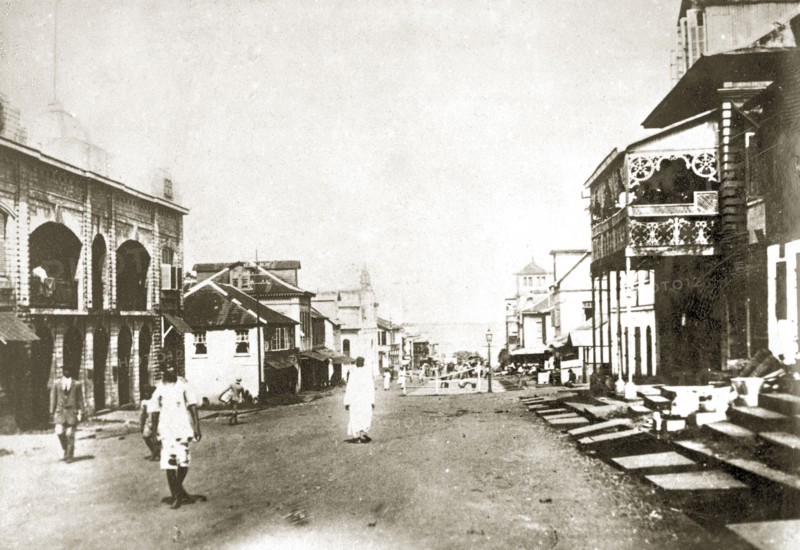 Sierra Leone street scene. A handful of people mill about on a wide road in the centre of a large town. The road is flanked by buildings of different styles and leads down towards the sea. Sierra Leone, 8-13 December 1923. Sierra Leone, Western Africa, Africa.