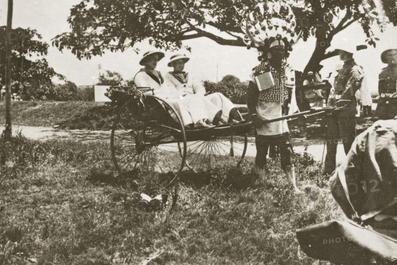 Rickshaw puller in Durban. A rickshaw puller wearing traditional costume and a flamboyantly feathered headdress gives a ride to two European men dressed in white naval uniforms. Durban, Natal (KwaZulu Natal), South Africa, 1-5 January 1924. Durban, KwaZulu Natal, South Africa, Southern Africa, Africa.
