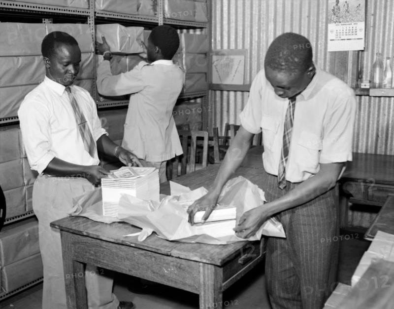 Packing books for dispatch. Promotional shot for the East African Literature Bureau. Three African men wearing shirts and ties wrap bundles of new books in paper and shelve them ready for dispatch. Kenya, 25 September 1956. Kenya, Eastern Africa, Africa.