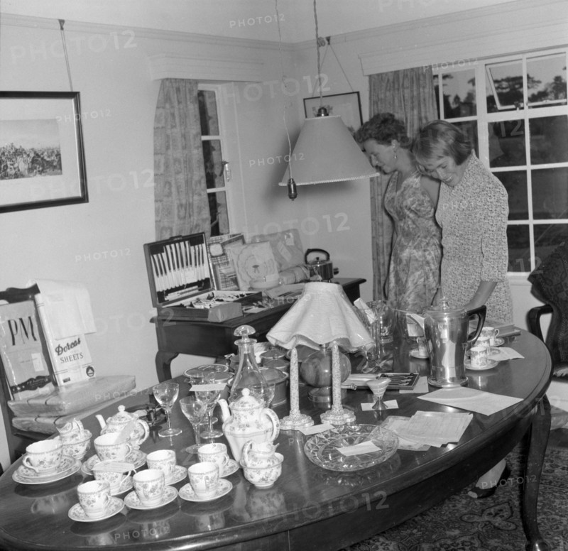 Inspecting the wedding presents. Two women examine wedding presents on display at the Patterson wedding. Amongst other items, the gifts include candlesticks, a tea set, a coffee jug, wine glasses, a decanter, a lamp shade, plates and a cutlery set. Kenya, 29 September 1956. Kenya, Eastern Africa, Africa.