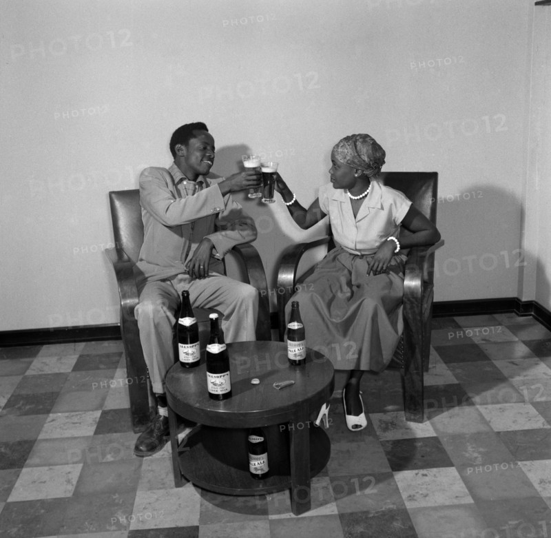 A toast to Allsopp's Pale Ale. Promotional shot for Allsop's Pale Ale. An African man and woman wearing Western clothes toast a drink to each other. Four bottles of Allsopp's Pale Ale and a bottle opener sit on the table in front of them. Kenya, 2 October 1956. Kenya, Eastern Africa, Africa.