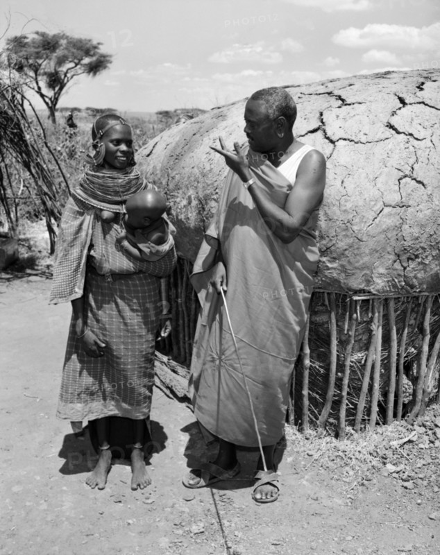 Lesanguniguni and his wife. A Samburu man identified as 'Lesanguriguri' chats to his young wife who holds a baby. Both wear traditional dress: her's decorated with striking, ornate jewellery. Wamba, Kenya, 13 October 1956. Wamba, Rift Valley, Kenya, Eastern Africa, Africa.