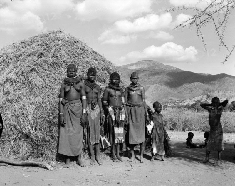 Turkana village girls. Four young Turkana women and children pictured outside a thatched village hut. They are semi-naked and wear traditional jewellery including heavily beaded necklaces, armlets and numerous hooped earrings. Wamba, Kenya, 13 October 1956. Wamba, Rift Valley, Kenya, Eastern Africa, Africa.
