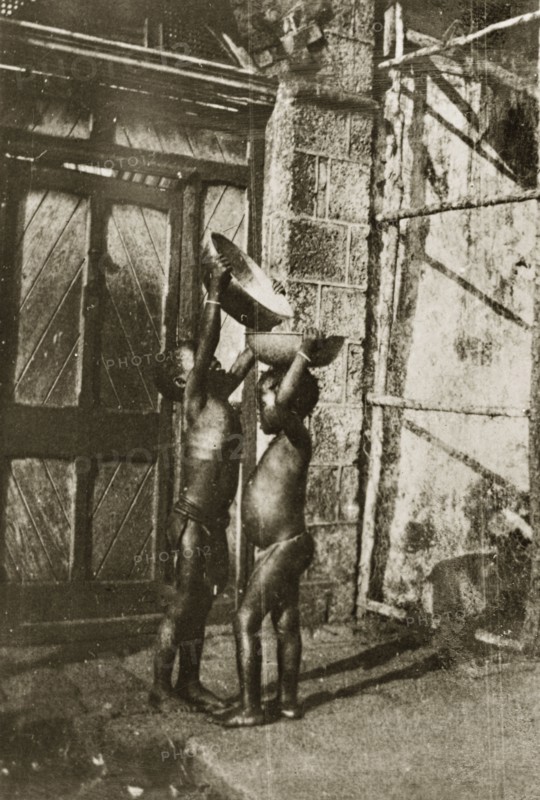 Young water carriers. A young child stretches up to pour water from one bowl into another balanced upon a friend's head. Ceylon (Sri Lanka), 27-31 January 1924. Sri Lanka, Southern Asia, Asia.