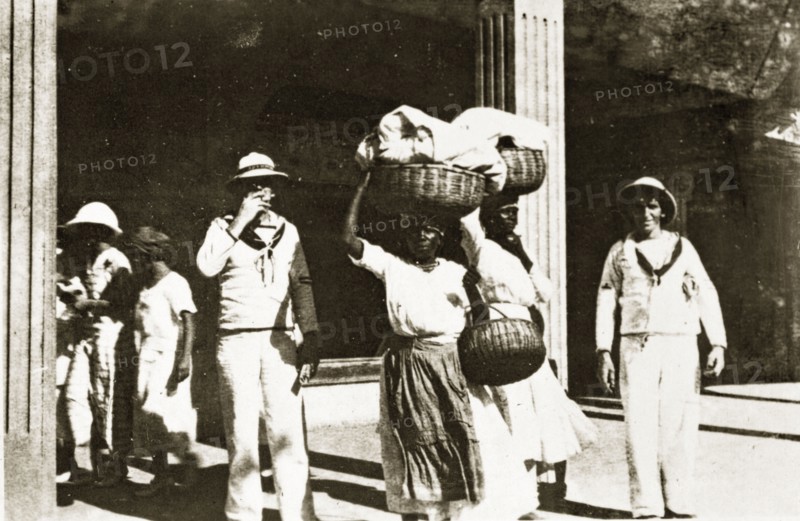 Jamaican street scene. Uniformed sailors from the British Special Service Squadron pose for the camera beside two Jamaican women carrying baskets of laundry on their heads. Kingston, Jamaica, 26-30 July 1924. Kingston, Kingston, Jamaica, Caribbean, North America .