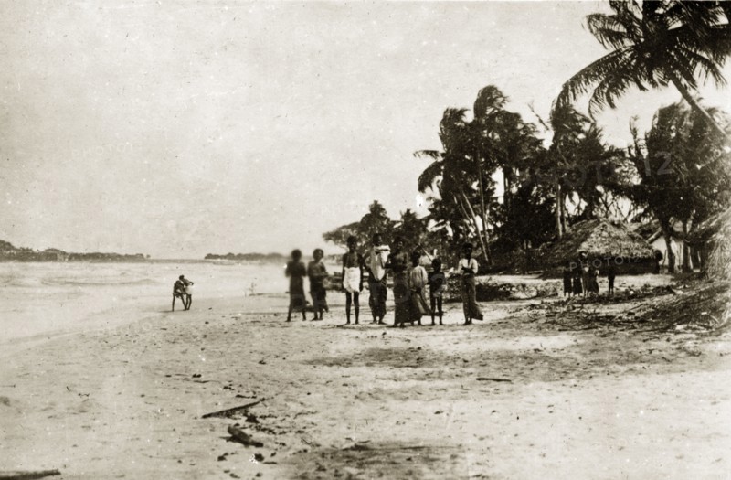 Ceylonian seashore. A group of Ceylonian people stand facing the camera on a beach. Thatched village huts and palm trees are visible in the background. Trincomoli, Ceylon (Sri Lanka), 27-31 January 1924. Trincomali, East (Sri Lanka), Sri Lanka, Southern Asia, Asia.