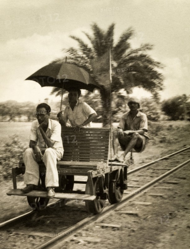 Railway hand cart. Three men sit aboard a small railway cart fitted with a wooden seat, an unusual vehicle reminiscent of a railway hand car. One of the men appears deep in contemplation, another holds up an umbrella. India, circa 1954. India, Southern Asia, Asia.