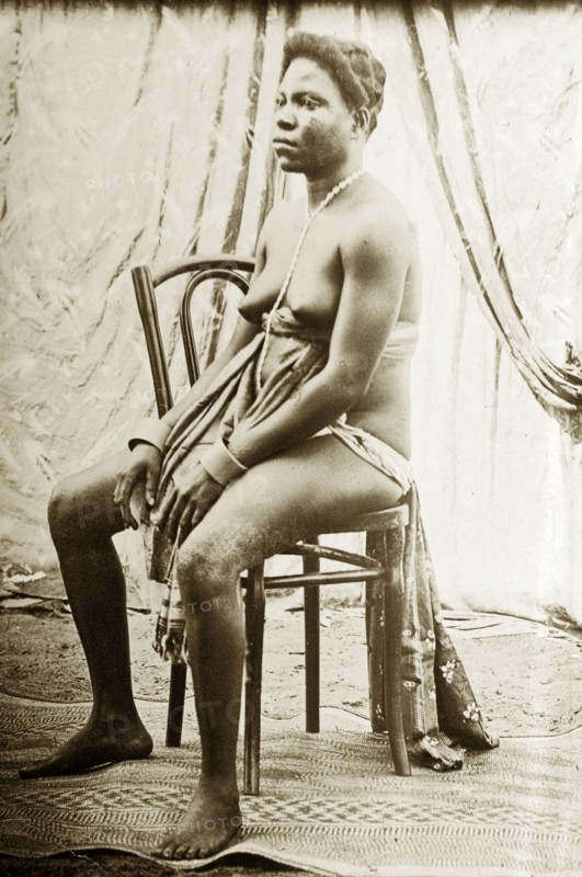Seated woman wearing apron and beads. Studio portrait of a seated woman, wearing an under-bust apron, waistcloth and simple jewellery. Western Africa, circa 1917., Western Africa, Africa.