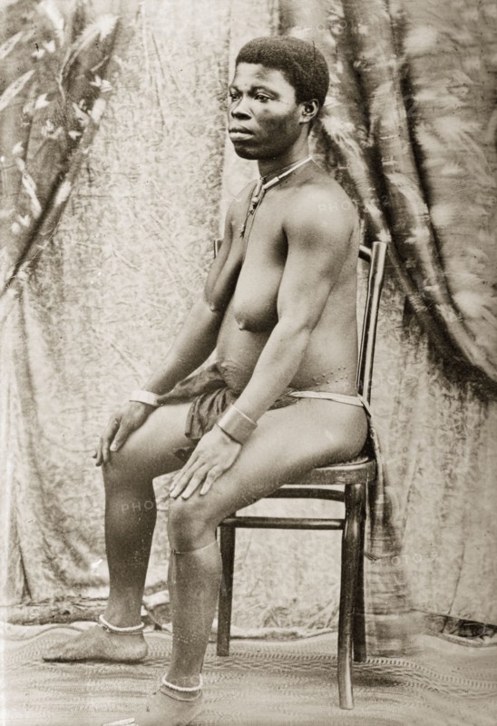 Seated woman with scarified stomach. Portrait of a seated woman, with scarified stomach and wearing an apron and back cloth around her waist and simple jewellery on her neck, wrists and ankles. Western Africa, circa 1917., Western Africa, Africa.