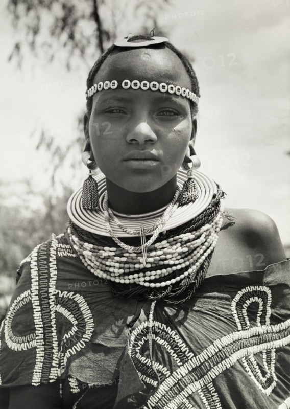 Young Maasai woman. Close-up portrait of a young Maasai woman wearing traditional jewellery including earrings, beaded necklaces and a beaded headband. Her wraparound robe is also adorned with beads sewn on in decorative patterns. Tanganyika Territory (Tanzania), circa 1960. Tanzania, Eastern Africa, Africa.