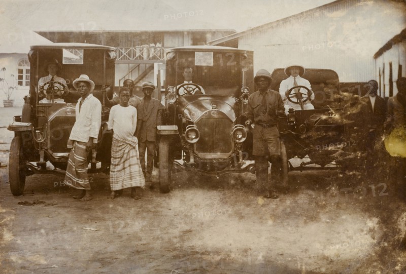 Men and cars, Western Africa. Three European drivers sit behind the wheels of their cars, surrounded by a several African men. Western Africa, circa 1920, Western Africa, Africa.