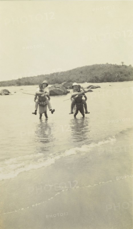 Out of water. Two European men wearing solatopi hats are carried above the water on the backs of two African men smoking cigarettes. Possibly Ghana, Western Africa, circa 1920., Western Africa, Africa.
