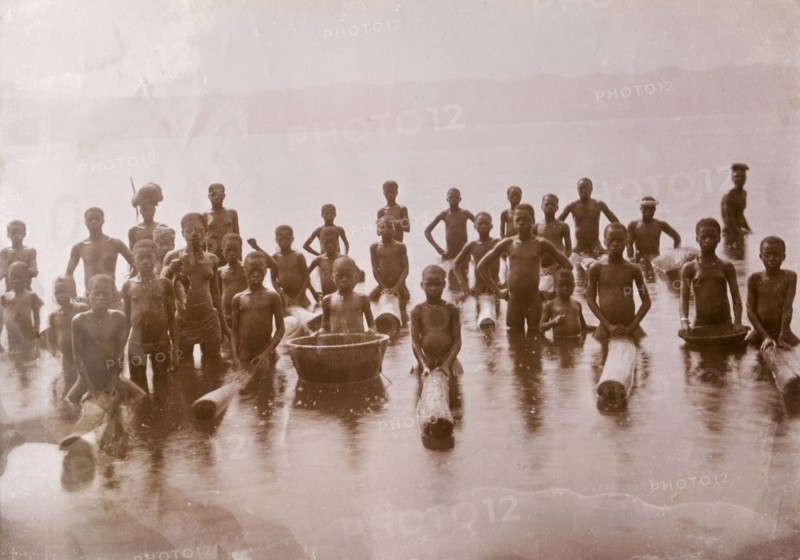 Children at lake shore, Gold Coast. A group of naked African people, mainly children, pose for the camera at a lake near Kumasi. Some of them sit astride floating logs, possibly a fishing aid or perhaps a recreational toy. Gold Coast (Ghana), circa 1920., Ashanti, Ghana, Western Africa, Africa.