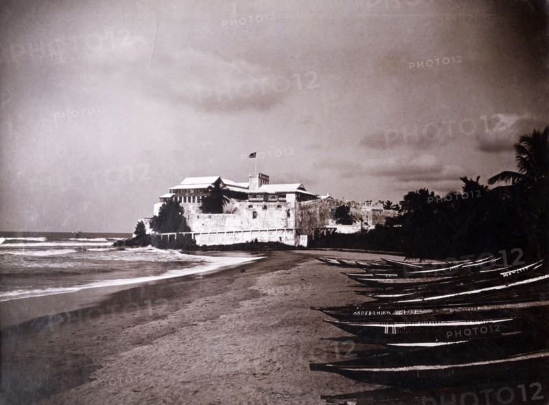 Christiansborg castle, Gold Coast. A line of canoes sit on a beach in front of Christiansborg castle or fort. Accra, Gold Coast (Ghana), circa 1920. Accra, East (Ghana), Ghana, Western Africa, Africa.
