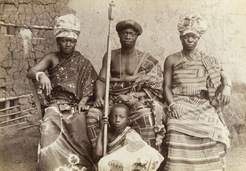The Mensa of Kumasi and his family. Outdoors portrait of the Mensa of Kumasi with his two wives and a son. He is shown here wearing a kente robe, as does his wife on the right. The wife on the left wears one or two wraps of tie-dye fabric, while the boy wears a robe of printed cotton. The child holds a long metal staff, possibly his father's staff of office. Kumasi, Gold Coast (Ghana), circa 1920. Kumasi, Ashanti, Ghana, Western Africa, Africa.