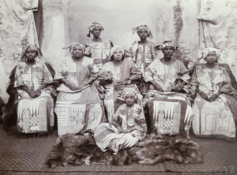 Eight women in indigo dresses. The women wear tailored gowns of dark cloth (probably indigo blue cotton) embroidered with geometrical patterns, suggesting that they are Hausa women from northern Nigeria. They also wear head cloths. Western Africa, circa 1918., Western Africa, Africa.