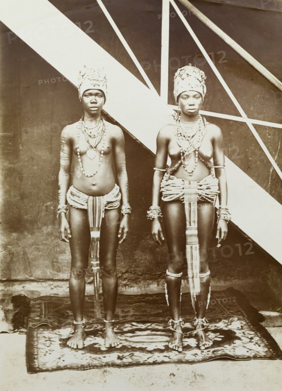 Two women in beaded dress, Gold Coast. Outdoors portrait of two young women wearing elaborate beaded and strip-cloth aprons, tall headdresses and heavy jewellery. Gold Coast (Ghana), circa 1920. Ghana, Western Africa, Africa.