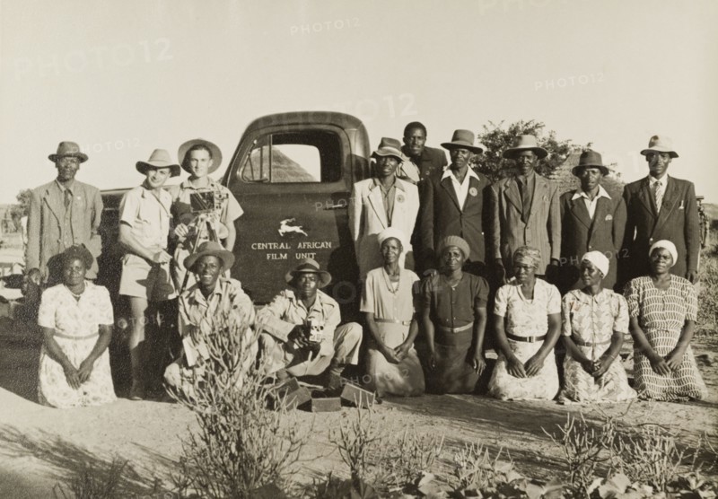 On location with the Central African Film Unit. Two European cameramen pose with the African cast and crew of a film being shot by the Central African Film Unit (CAFU). The group stand in front of a company vehicle on location in the countryside. Probably Southern Rhodesia (Zimbabwe), circa 1954. Zimbabwe, Southern Africa, Africa.