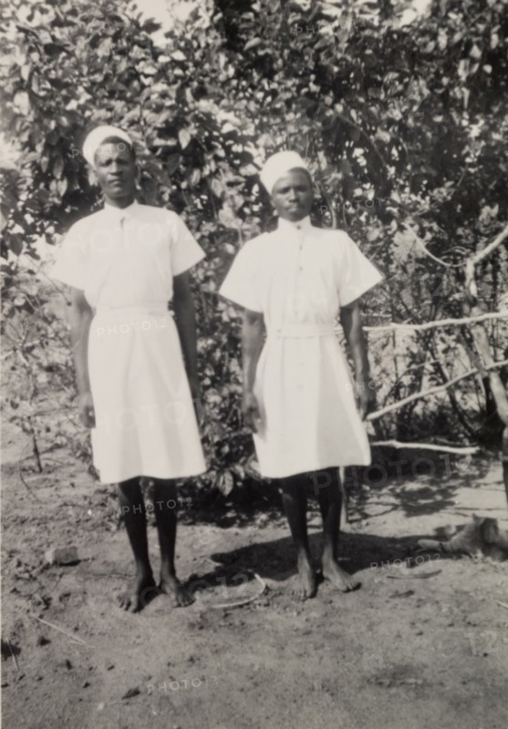 Cigarette and Timothy'. Two domestic servants, identified by an original caption as 'Cigarette and Timothy', pose for the camera. Their pristine uniform consists of a knee-length white dress, belted at the waist and worn with a starched white hat. Northern Rhodesia (Zambia), circa 1952. Zambia, Southern Africa, Africa.