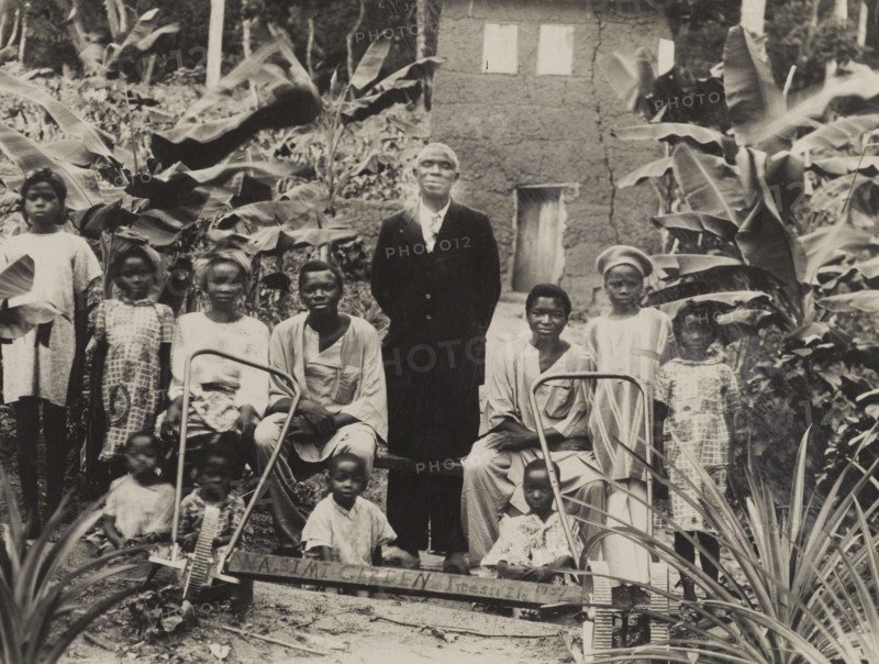 Children in 'Wasimi Garden'. Children gather around an adult male for a group photograph in 'Wasimi Garden', the name indicated by a handmade sign. Two hand-operated tilling machines sit in the foreground. Imesi-Ile, Nigeria, 1951. Imesi-Ile, Osun, Nigeria, Western Africa, Africa.