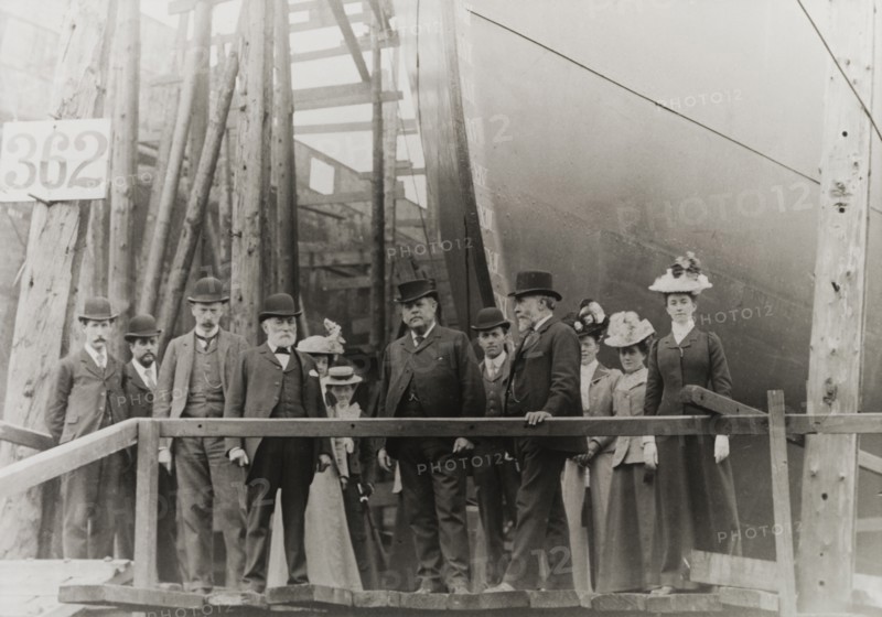 Sir Alfred Holt at a dockyard. A group of formally dressed men and women pose for the camera beside the bow of a large ship in dry dock. Amongst the group is Sir Alfred Holt (fourth from right), founder of the Blue Funnel shipping line. United Kingdom, circa 1890. England (United Kingdom), Western Europe, Europe .