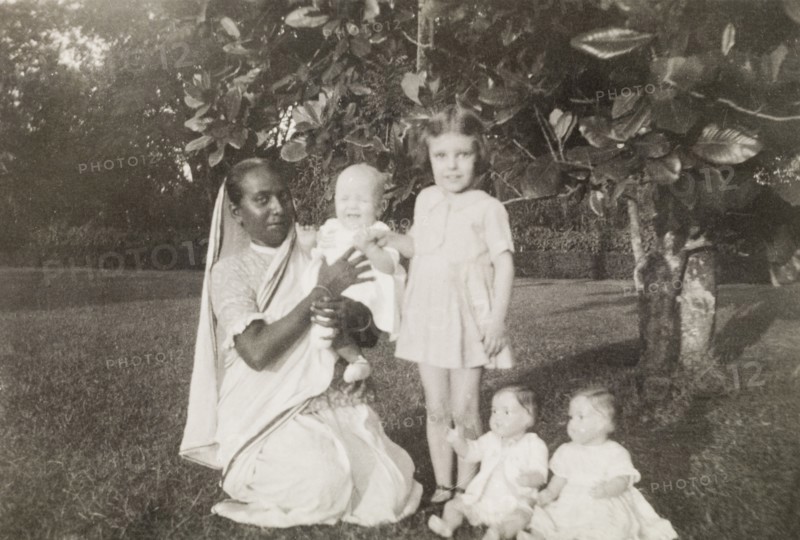 Ayah with children. An Indian ayah (nursemaid) poses beside her British charges, a young girl and a baby, in the garden of a colonial residence. Two toy dolls are propped up for the camera in the foreground. Calcutta (Kolkata), India, October 1932. Kolkata, West Bengal, India, Southern Asia, Asia.