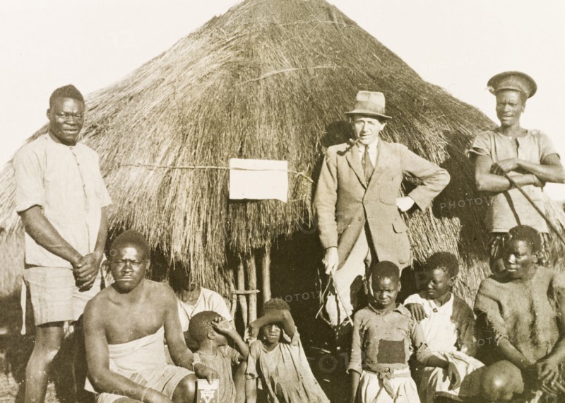 Portrait of workers and children at Balloch Farm. Portrait of a group of male African farm workers, assembled outdoors beside a round, thatched hut at Balloch Farm. They pose with four children and a European man, probably an overseer, who wears a suit and smokes a pipe. Near Gwelo, Southern Rhodesia (near Gweru, Zimbabwe), circa 1930. Gweru, Midlands, Zimbabwe, Southern Africa, Africa.