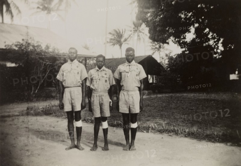 Three Kenyan scouts. Three Kenyan scouts pose for the camera outdoors at an inspection of scouts and guides by Sir Charles Mortimer, the Commissioner for Lands. Each boy wears scout uniform and what appears to be a bandage around his left knee. Coast Province, Kenya, circa 1950., Coast, Kenya, Eastern Africa, Africa.