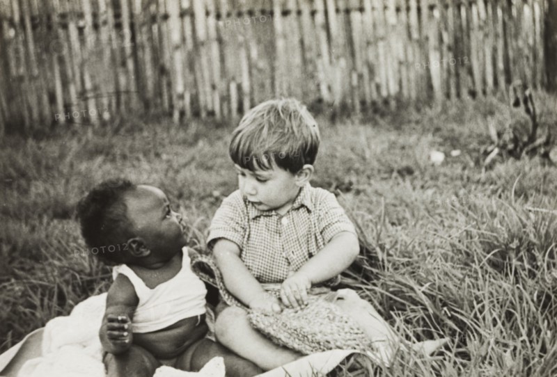 Anthony Lewis befriends an African baby. Anthony Lewis, a youngest son of English Methodist missionary, Reverend Ian Lewis, sits beside an African baby on a patch of grass outside the Lewis family home. Limuru, Kenya, circa 1956. Limuru, Central (Kenya), Kenya, Eastern Africa, Africa.