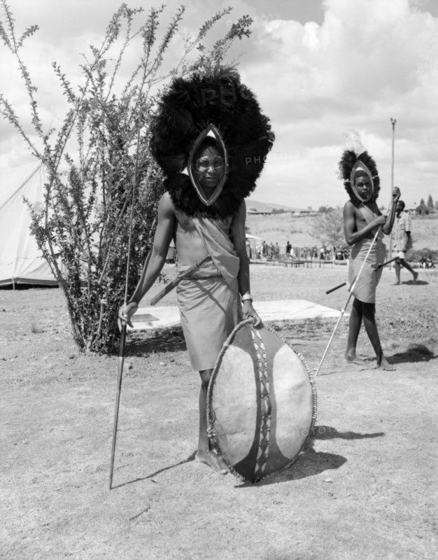 Portrait of a male Wakamba dancer. Portrait of a male Wakamba dancer wearing a large, mane-like headdress made from black feathers for Princess Margaret's visit. His face is painted and he holds a patterned shield and spear. Machakos, Kenya, 22 October 1956. Machakos, East (Kenya), Kenya, Eastern Africa, Africa.