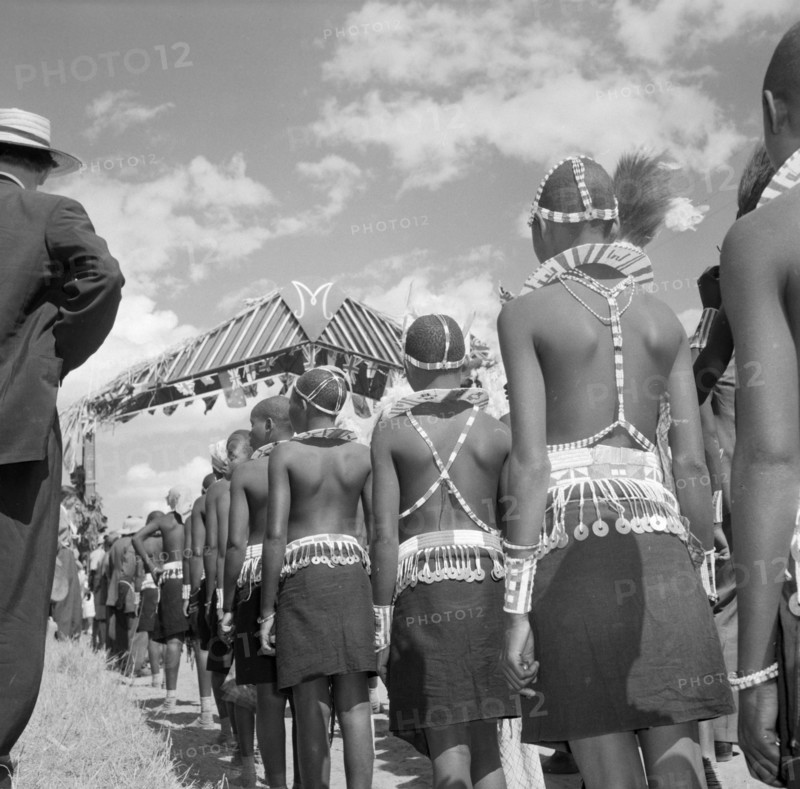 Wakamba costume from behind. Wakamba dancers stand in line with their backs to the camera during Princess Margaret's visit. Their ceremonial dress consists of elaborately beaded necklaces, headbands and belts. Machakos, Kenya, 22 October 1956. Machakos, East (Kenya), Kenya, Eastern Africa, Africa.