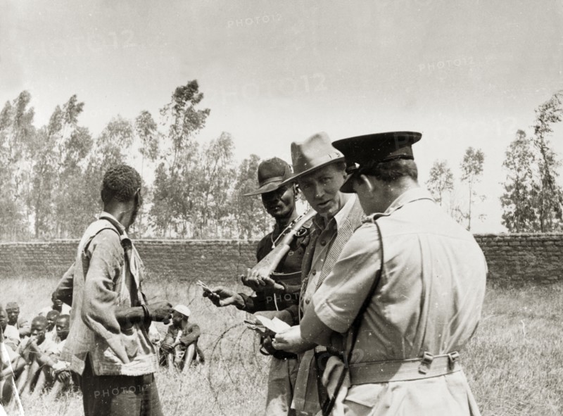Searching for Mau Mau suspects. A farmer and military officers question a Kikuyu farm labourer. Behind him more Kikuyu sit on the ground surrounded by coils of barbed wire. Kenya, circa 1953. Kenya, Eastern Africa, Africa.