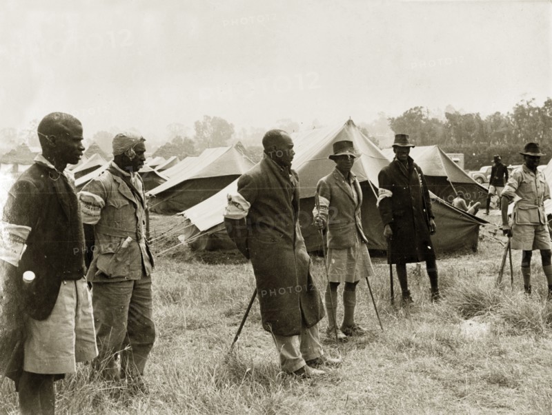 Kikuyu Home Guards in camp. Six Kikuyu Home Guards, dressed in makeshift uniforms and armed with spears, stand in front of rows of substantial tents, presumably a temporary home guard post. Kenya, circa 1952. Kenya, Eastern Africa, Africa.