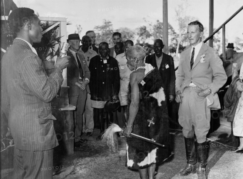 Maina arap Chumo at a function. Maina arap Chumo, a Kikuyu elder dressed in a cowhide wrap and carrying a whisk, arrives or speaks at an official or settler gathering in Nakuru. A radio interviewer stands nearby. Nakuru, Kenya circa 1953. Nakuru, Rift Valley, Kenya, Eastern Africa, Africa.