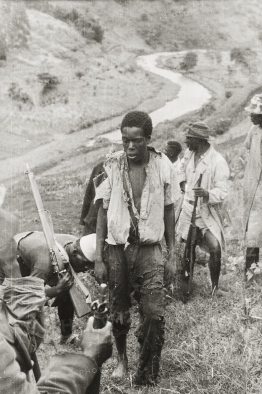 Captured Mau Mau suspect. A captured Mau Mau suspect, wearing a tattered shirt and trousers, stands on a hillside surrounded by heavily armed members of the Kikuyu Home Guard. Kenya, circa 1952. Kenya, Eastern Africa, Africa.