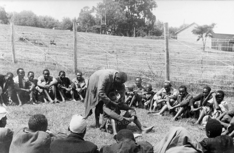 Government de-oathing ceremony. A Mau Mau detainee sitting in the centre of a circle of men in a fenced compound is 'purified' of the Mau Mau oath. Kenya, circa 1954. Kenya, Eastern Africa, Africa.