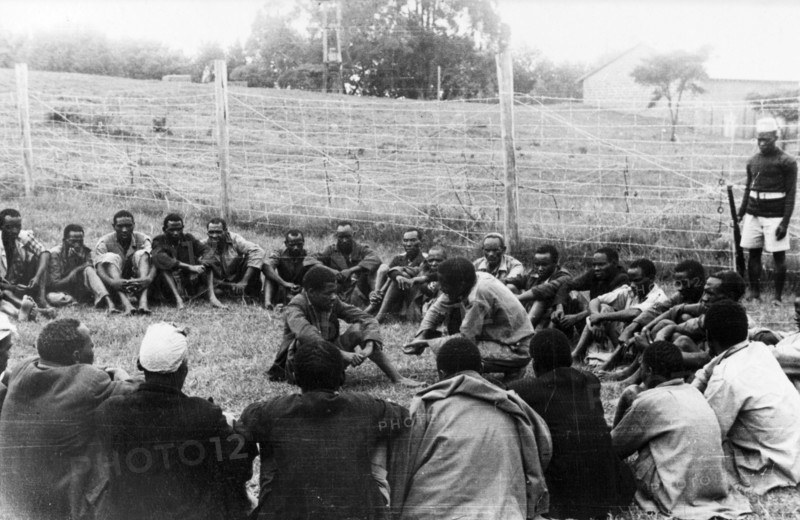Government de-oathing ceremony. A Mau Mau detainee sitting in the centre of a circle of men in a fenced compound is prompted to confess to having taken the Mau Mau oath. Kenya, circa 1954. Kenya, Eastern Africa, Africa.